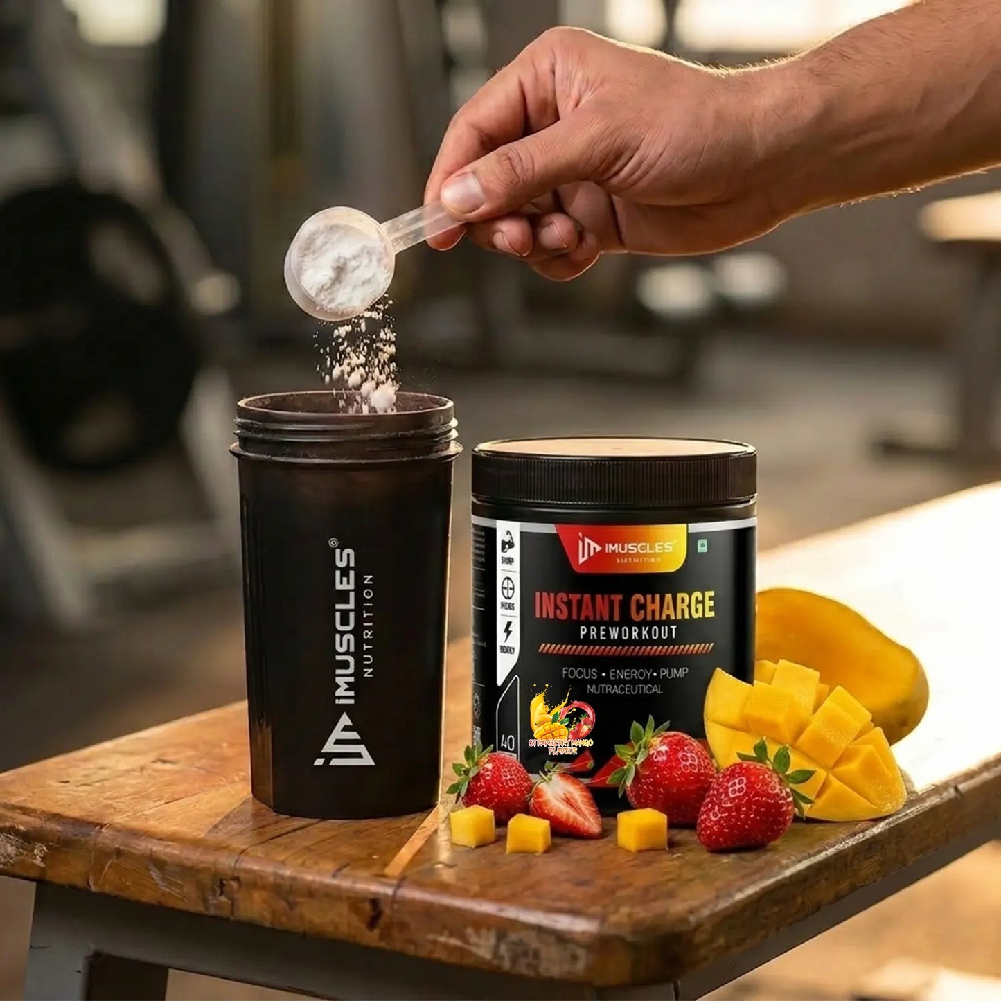 A square digital product promotional image titled "Strawberry Mango 7.jpg" showing a close-up action shot on a wooden gym bench. A hand holds a clear scoop, pouring white pre-workout powder into an open black iMuscles Nutrition shaker bottle on the left. Next to it stands a black iMuscles Nutrition "INSTANT CHARGE PRE-WORKOUT" jar. The scene is decorated with fresh, sliced strawberries and neatly cubed mango pieces, with a blurred gym background.