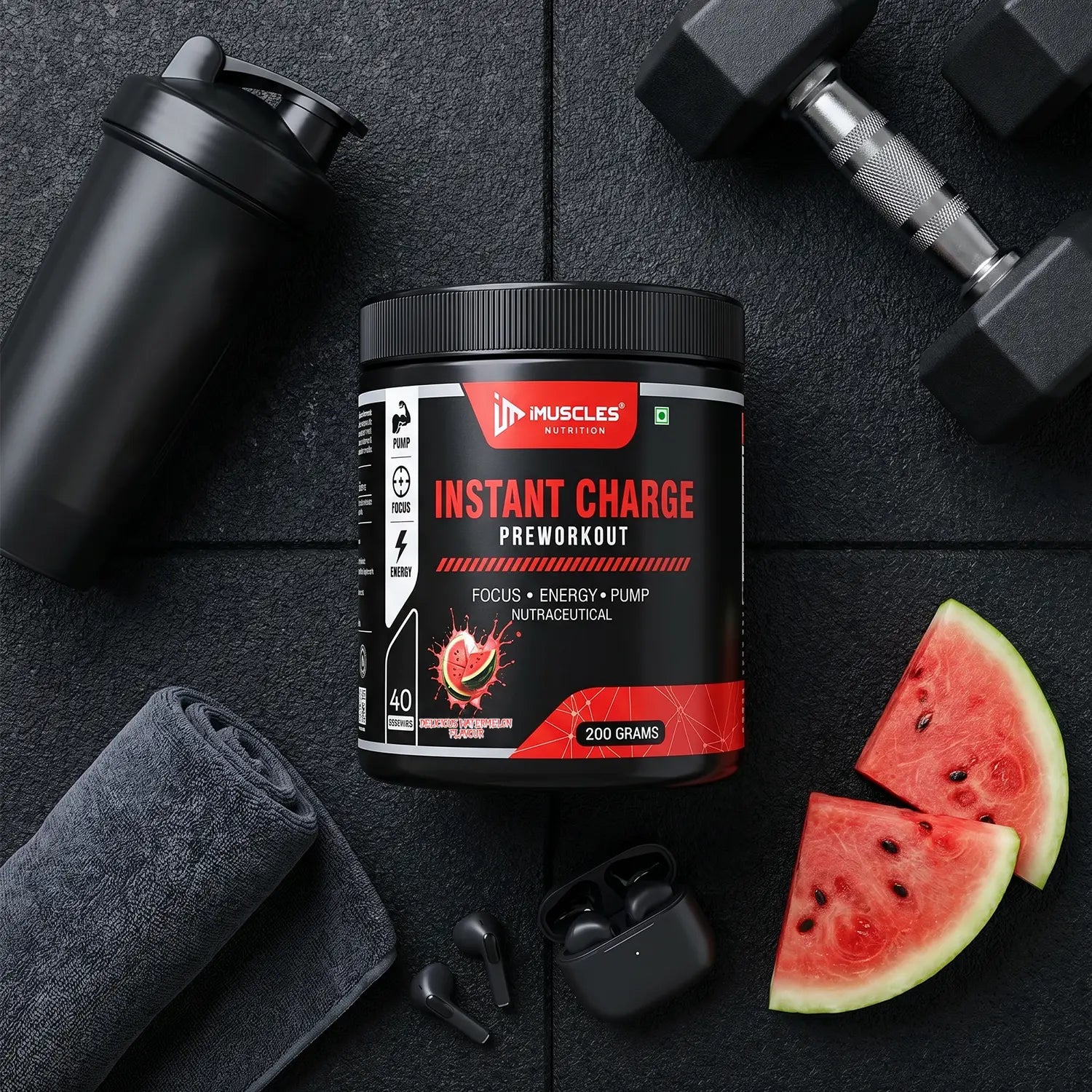 Top-down gym flat-lay featuring iMUSCLES NUTRITION Instant Charge Pre-Workout in Delicious Watermelon flavor. The 200g container is arranged on a black textured gym mat alongside a matte black shaker bottle, a pair of hex dumbbells, a rolled-up grey workout towel, wireless black earbuds with their charging case, and fresh watermelon slices.