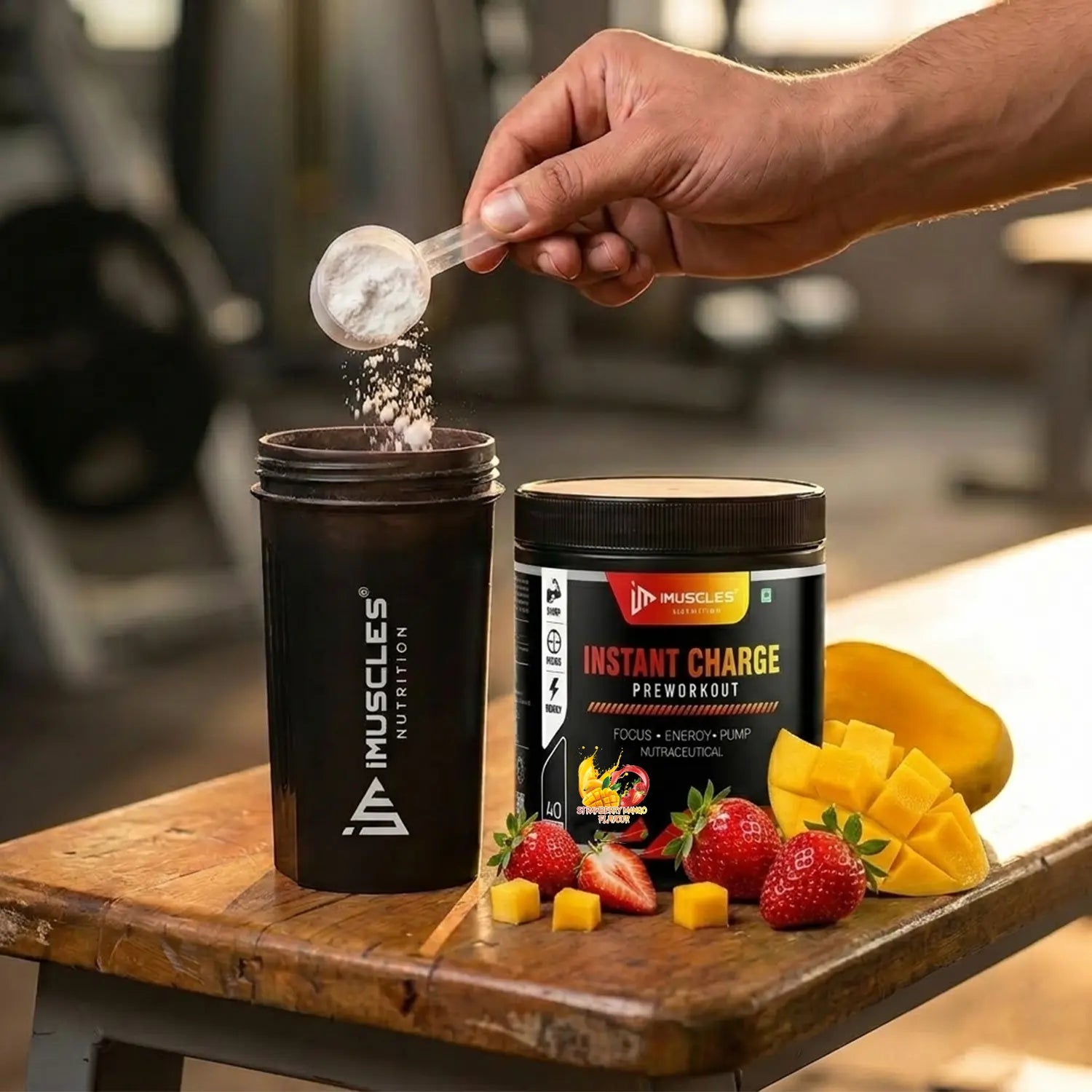 A square digital product promotional image titled "Strawberry Mango 7.jpg" showing a close-up action shot on a wooden gym bench. A hand holds a clear scoop, pouring white pre-workout powder into an open black iMuscles Nutrition shaker bottle on the left. Next to it stands a black iMuscles Nutrition "INSTANT CHARGE PRE-WORKOUT" jar. The scene is decorated with fresh, sliced strawberries and neatly cubed mango pieces, with a blurred gym background.
